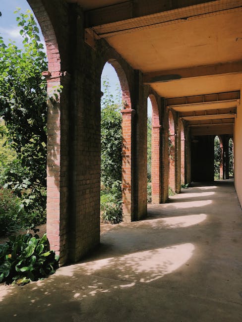 A covered outdoor walkway featuring a series of brick arches supported by brick columns, casting shadows onto the smooth concrete floor. To the left, lush green foliage with broad leaves and dense shr