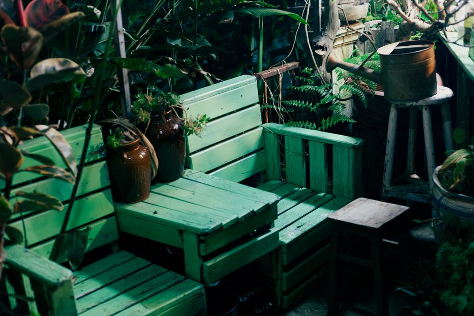 A cozy garden seating area featuring rustic wooden furniture painted in a soft green hue, including a bench with a slatted backrest and a matching armchair. The scene is lush with greenery, comprising